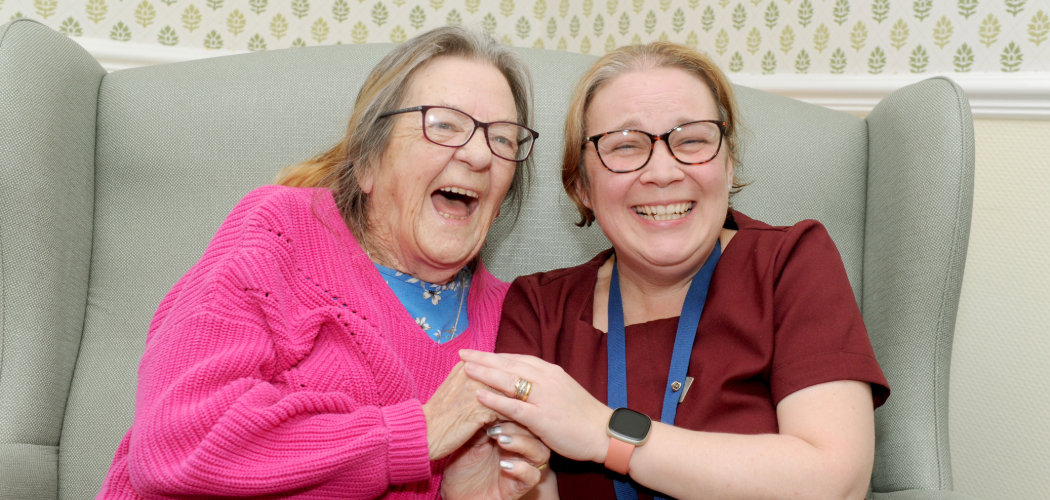 Two people are sitting closely together on a light green armchair. The person on the left is wearing a bright pink sweater over a blue top, while the person on the right is wearing a dark red shirt with a blue lanyard and an orange wristwatch. They are holding hands and are laughing. The background features wallpaper with small green leaf patterns and white trim.