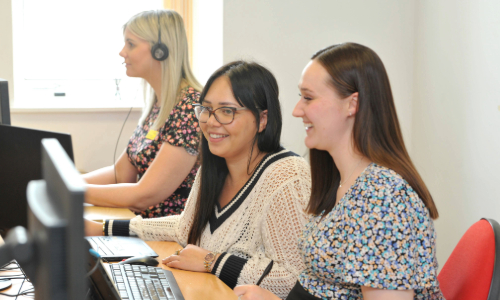 Three individuals are seated at a desk, working on desktop computers. The person on the left is wearing a headset and has blonde hair, while the two individuals on the right have dark hair and are focused on their screens. The background shows a window with natural light coming in