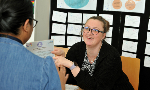 A member of the patient experience team is showing a member of the public some information about their services. She is smiling and wears a black cardigan, patterned shirt and glasses