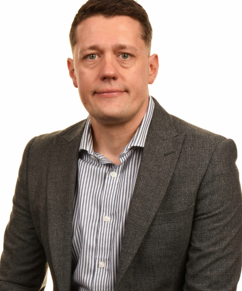 Headshot of James Fleet, he is facing the camera and smiling. He is wearing a blue and white striped shirt and grey suit jacket