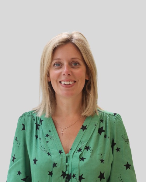 Head and shoulders shot of Laura looking into the camera directly and smiling. She has blonde hair and is wearing a green shirt with black stars